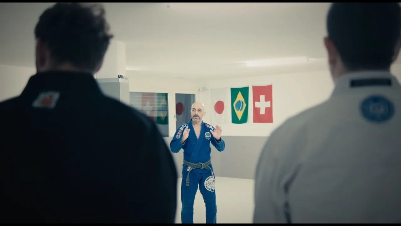 Standbild aus dem Video: Trainer im blauen Kimono spricht bei der Gürtelverleihung zur Gruppe, dahinter die Flaggen von Japan, Brasilien und der Schweiz