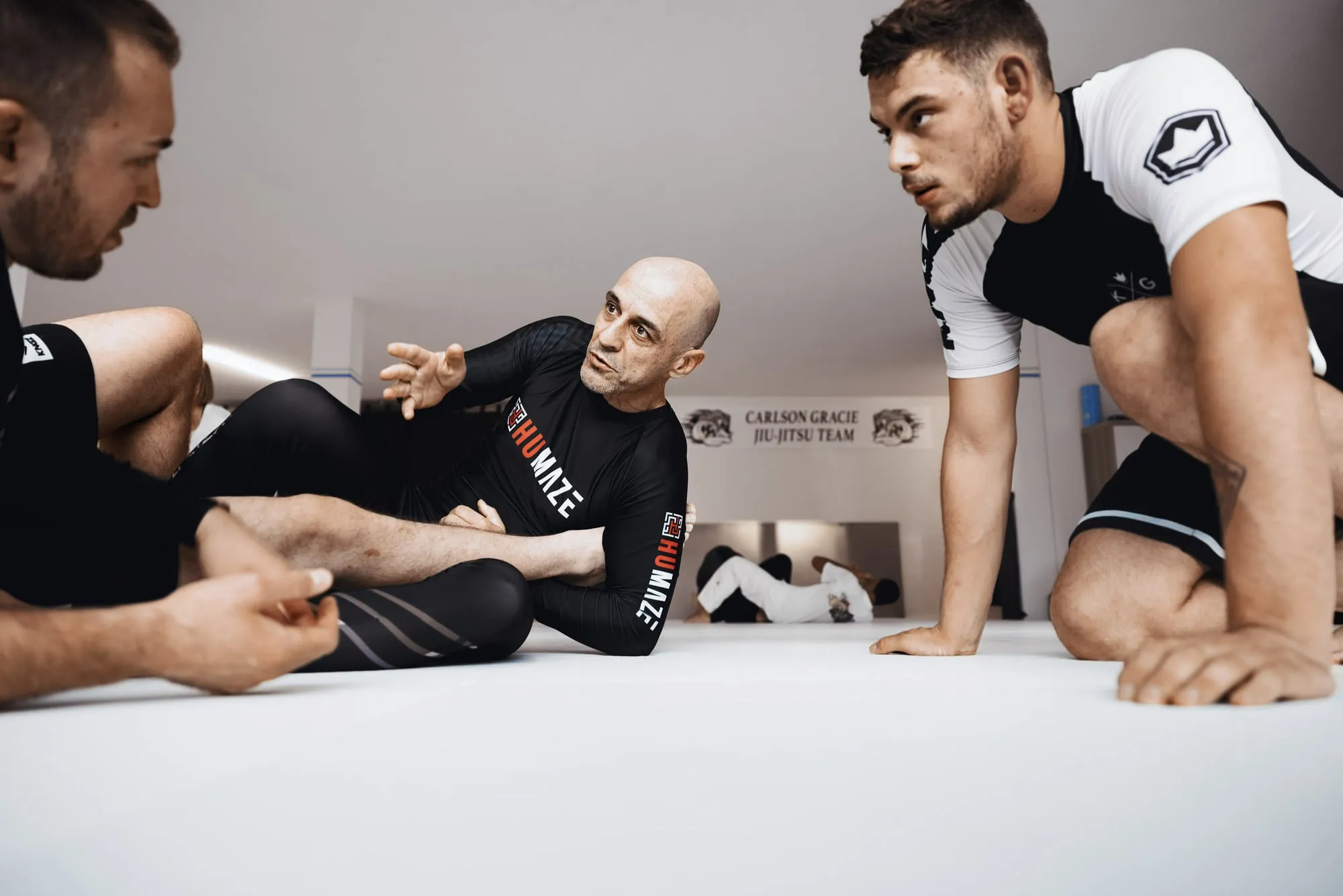 Trainer sitzt auf der Matte und erklärt zwei Trainierenden den nächsten Schritt