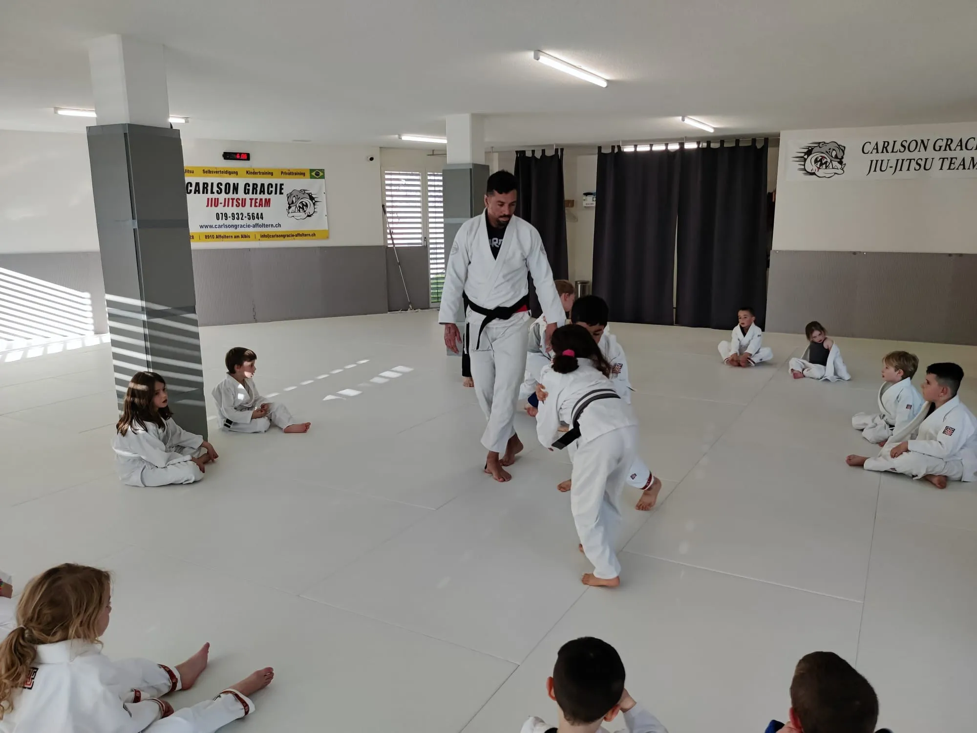 Kindertraining in der Carlson Gracie Academy Affoltern: Der Trainer begleitet zwei Kinder beim Üben, die übrigen Kinder sitzen am Mattenrand
